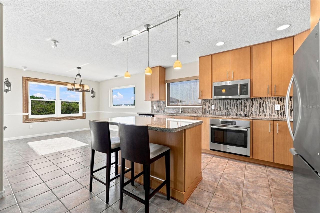 Photo 14 of 20: Kitchen — Renovated kitchen showing granite island, stainless microwave and wall oven, and glass mosaic tile backsplash with natural light from multiple windows.
