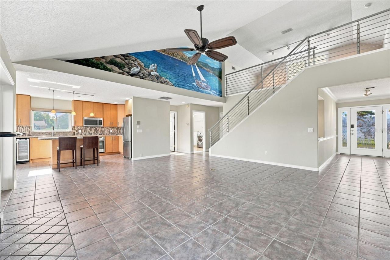 Photo 7 of 20: Great room — Open-concept great room with tile flooring, custom hand-painted coastal mural along the vaulted ceiling, staircase with cable railing, and kitchen visible beyond.