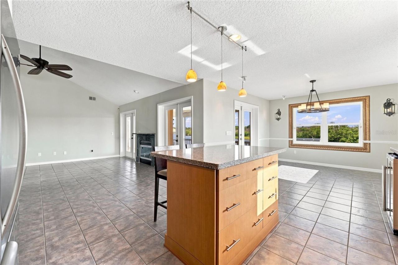 Photo 17 of 20: Kitchen and dining area — Open kitchen and breakfast area with granite island seating, chandelier, gas fireplace visible beyond, and framed picture window with canal views.