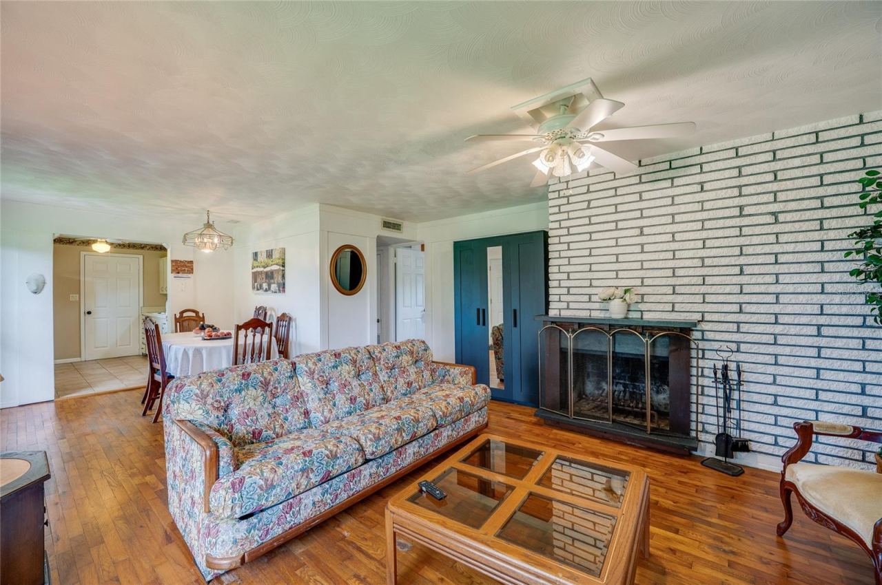 Photo 8 of 20: Open-concept main living space featuring rich wood flooring, a central ceiling fan, and a painted brick fireplace. The seamless floor plan connects adjacent living zones.