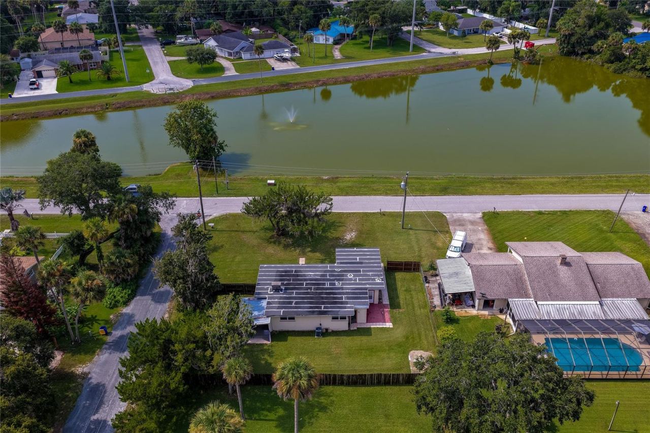 Photo 4 of 20: High-angle aerial view of the single-story home situated directly across from a peaceful neighborhood lake with a central water fountain. The spacious property features a grassy yard surrounded by mature palm trees and lush local vegetation.