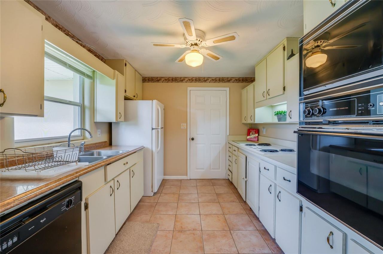 Photo 12 of 20: Functional galley-style kitchen featuring tile flooring, classic cabinetry, a double sink, and a built-in wall oven and microwave combo.