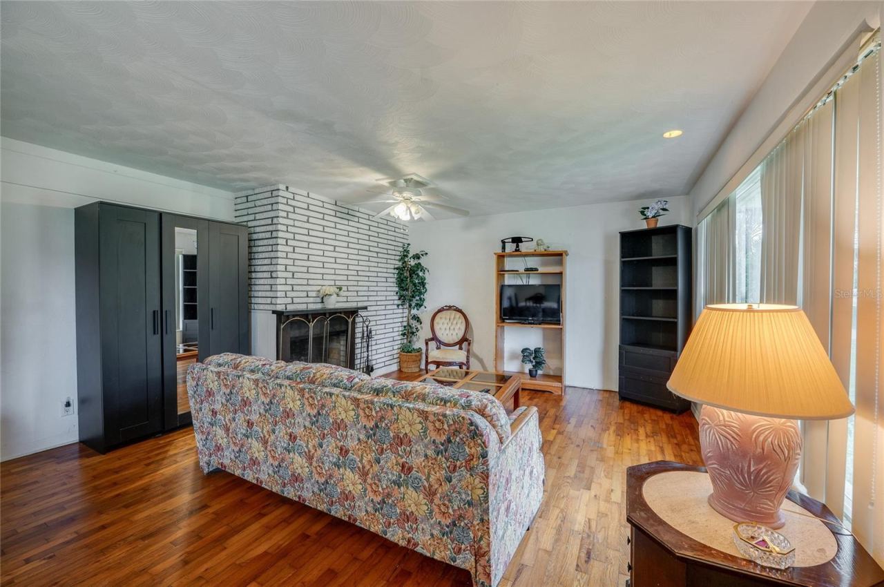 Photo 6 of 20: Warm living area featuring rich hardwood flooring, and a painted brick fireplace. Large windows bring in ample natural light to illuminate the inviting space.
