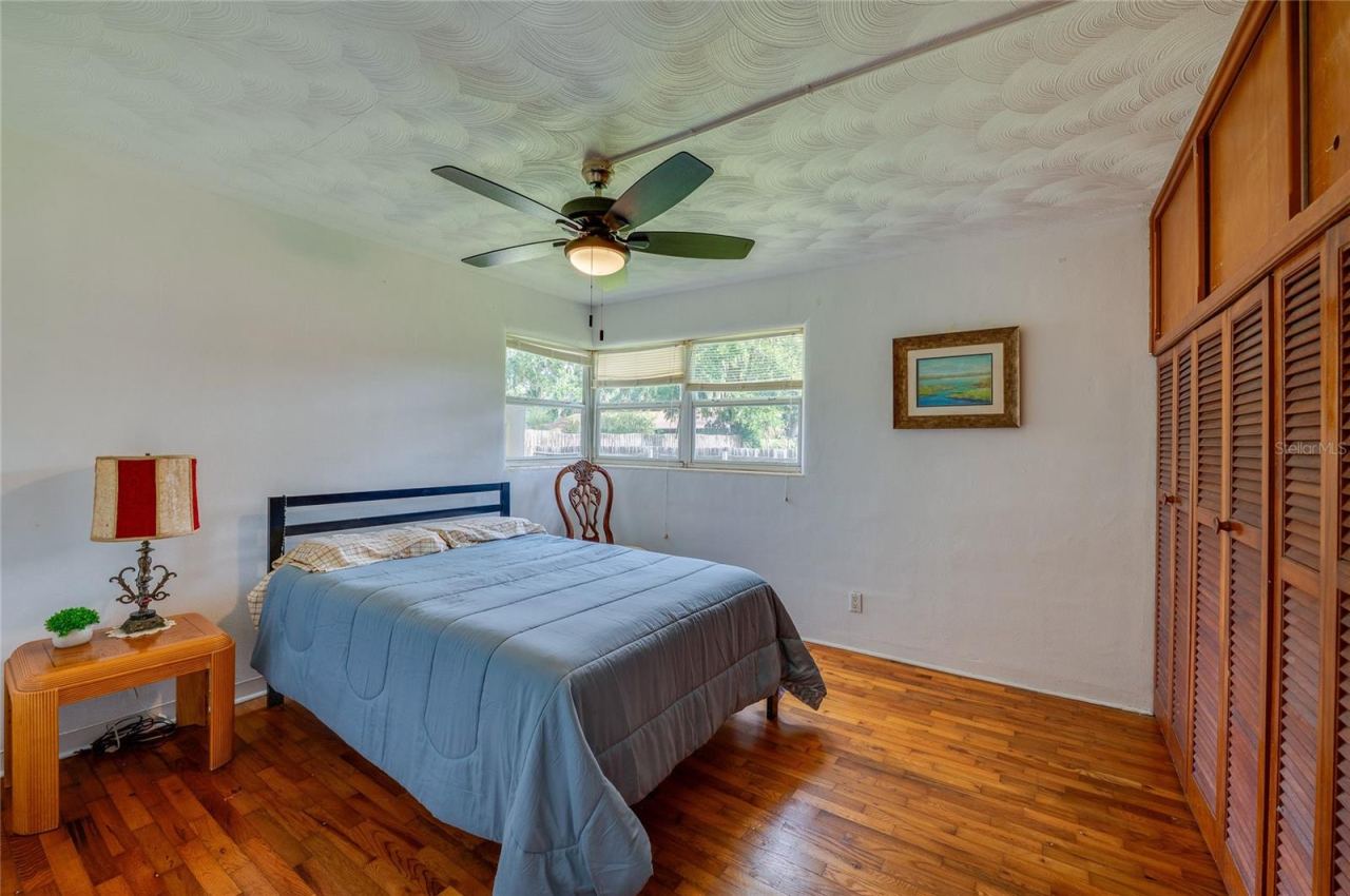 Photo 20 of 20: Bedroom area defined by warm hardwood flooring, a textured white ceiling with a fan, and full-height closet doors. Corner windows draw in natural light to brighten the layout.