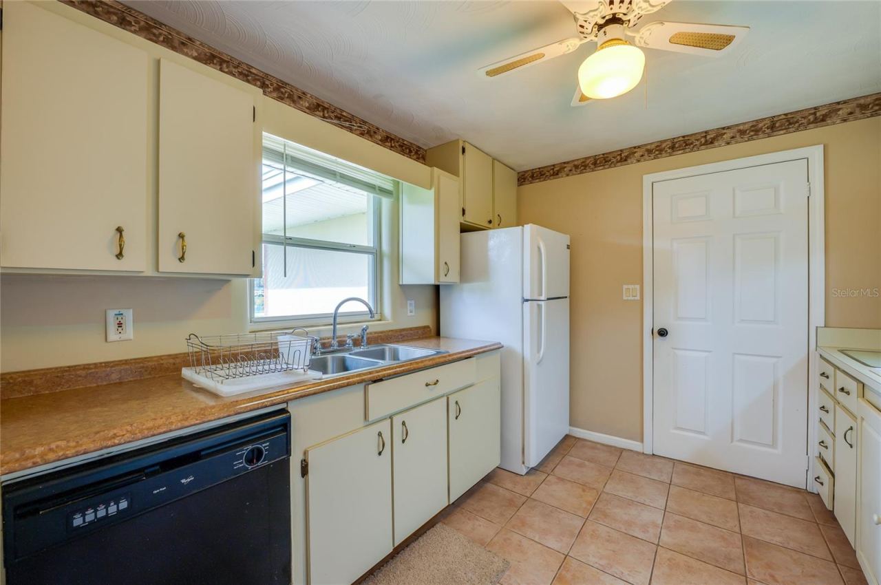 Photo 13 of 20: Kitchen work area showing a double basin sink, dishwasher, refrigerator, and neutral tiled flooring beneath an overhead ceiling fan.