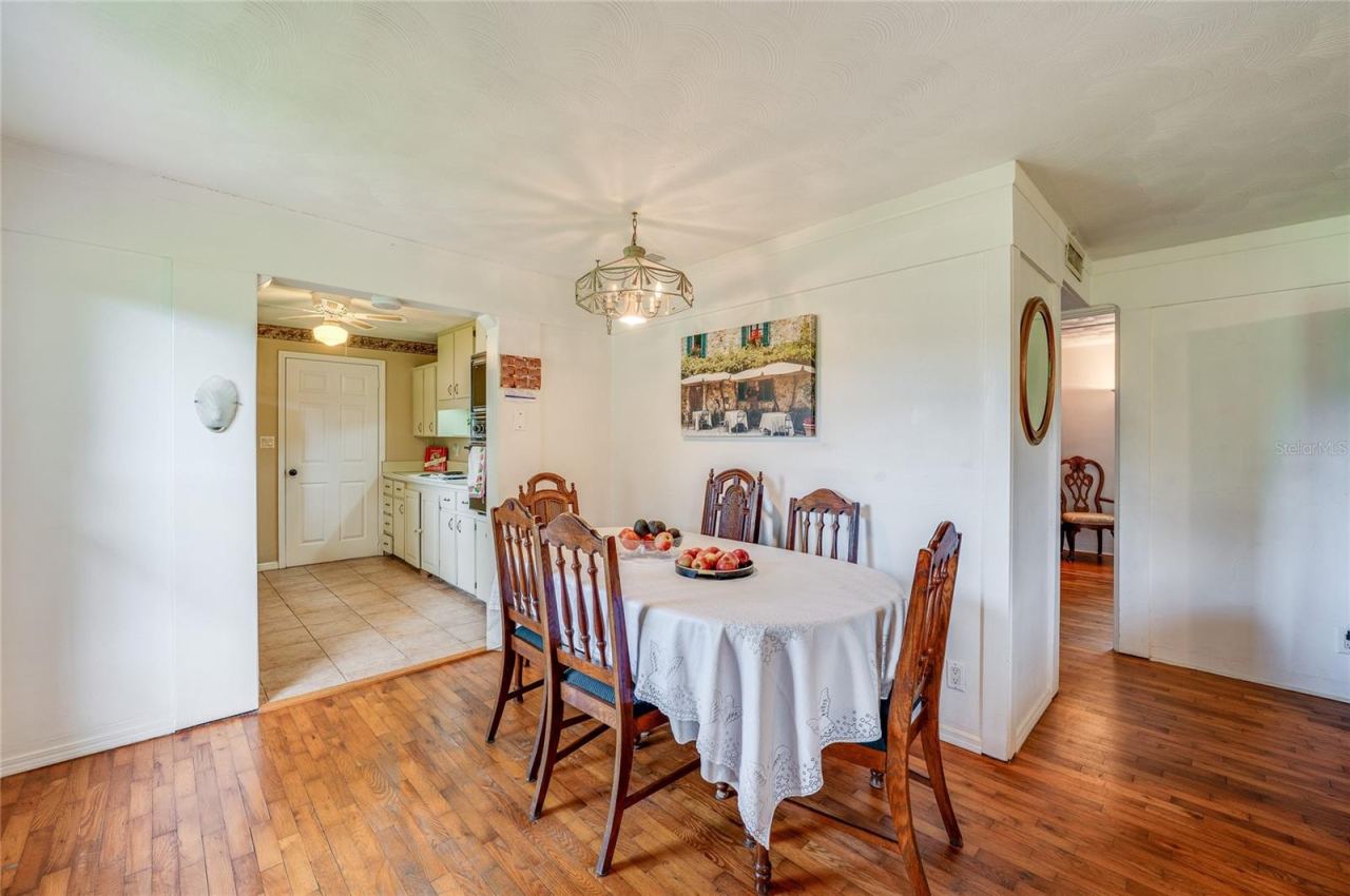 Photo 10 of 20: Dining area transitioning smoothly into the kitchen, complete with wood-look flooring, light walls, and tiled kitchen flooring beyond. A hanging overhead light fixture anchors the room while open doorways maintain an airy flow.