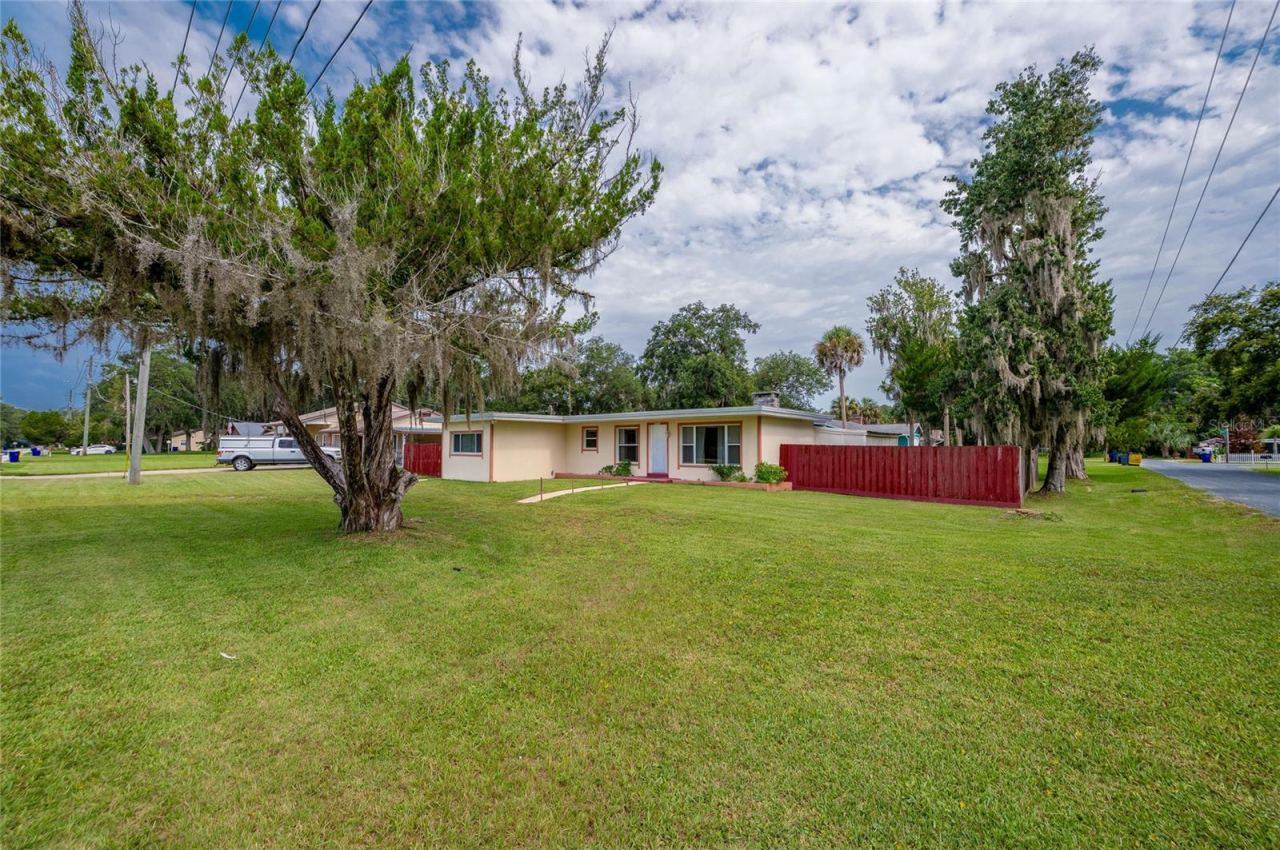 Photo 2 of 20: Single-story home framed by expansive green lawns, mature shade trees draped in Spanish moss, and a crimson perimeter fence.