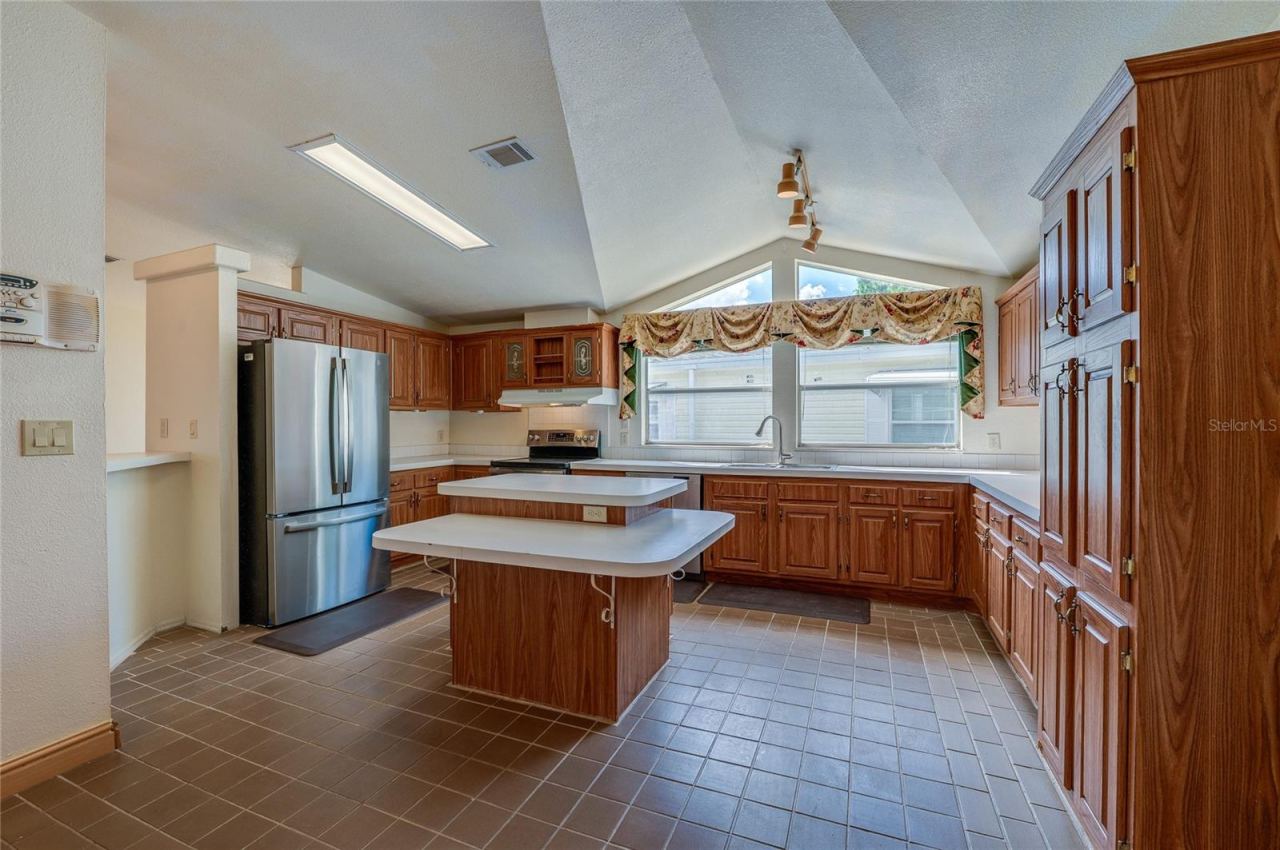 Photo 9 of 20: Spacious kitchen features abundant cabinetry, extensive counter space, a center island, stainless steel refrigerator, and expansive windows beneath a vaulted ceiling.