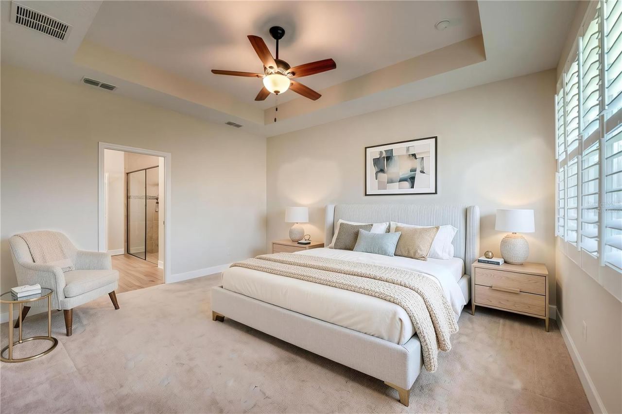 Photo 19 of 20: Virtually Staged - Spacious primary bedroom featuring neutral carpeting, a high tray ceiling with a ceiling fan, and a doorway leading into the en-suite bathroom.