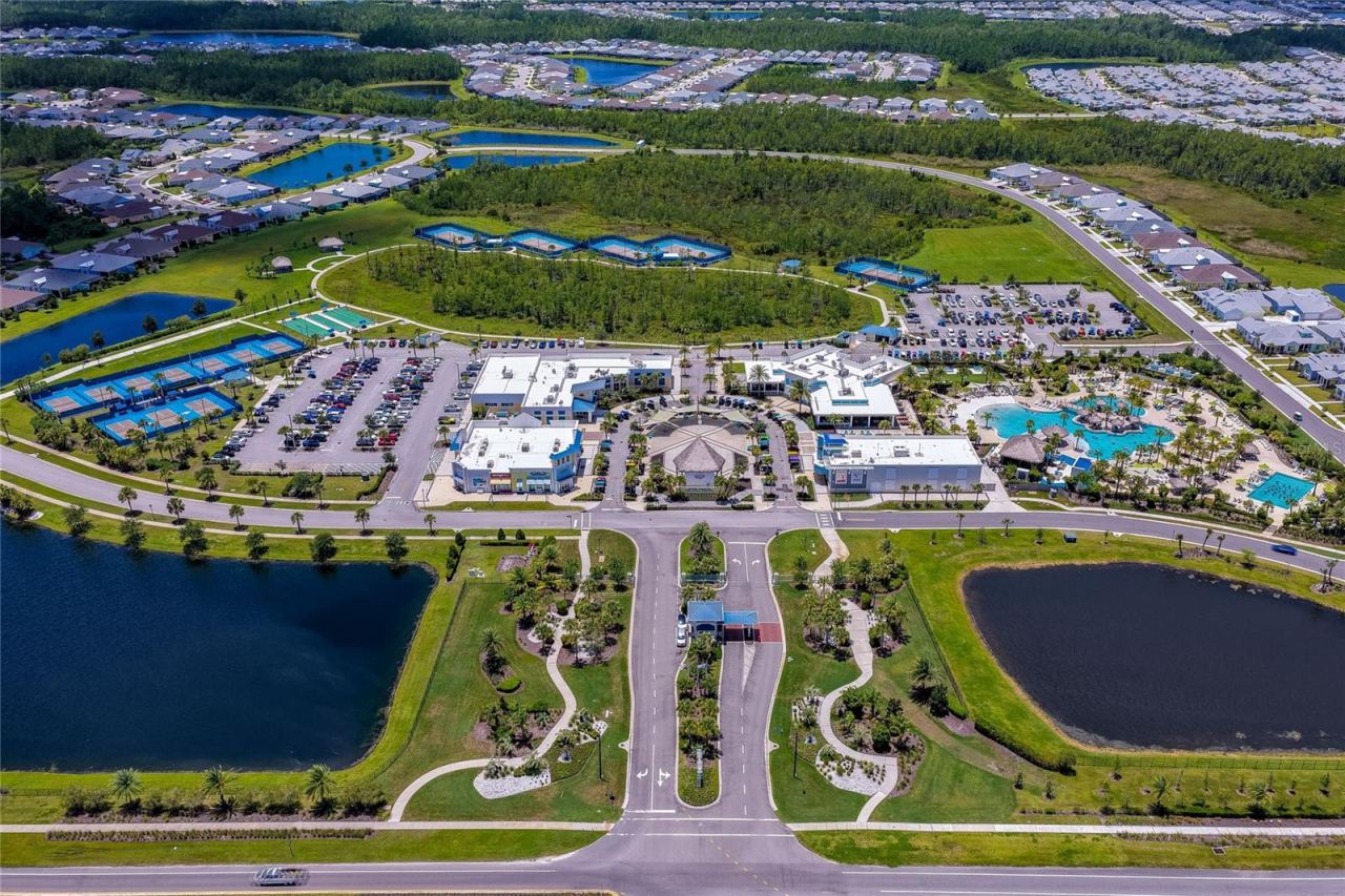 Photo 6 of 20: Birds-eye view of the expansive community clubhouse complex, featuring a resort-style swimming pool, numerous sports courts, and convenient parking facilities bordered by scenic lakes.