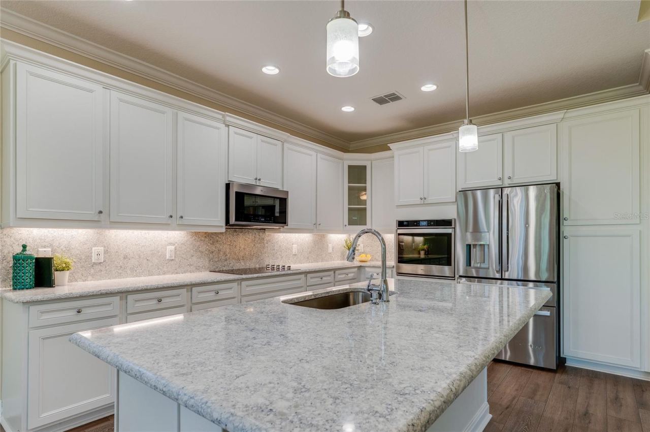 Photo 10 of 20: Elegant kitchen showing a spacious granite-topped island with an under-mount sink, premium white cabinetry, and modern stainless steel appliances. Under-cabinet lighting highlights the granite backsplash and countertops.