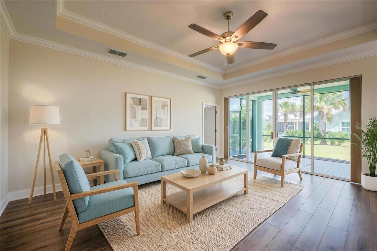 Photo 15 of 20: Virtually Staged - Bright, open living area featuring warm luxury vinyl flooring, neutral walls, and a tray ceiling with crown molding and a ceiling fan. A large sliding glass door opens to a screened-in patio with views of tropical landscaping.
