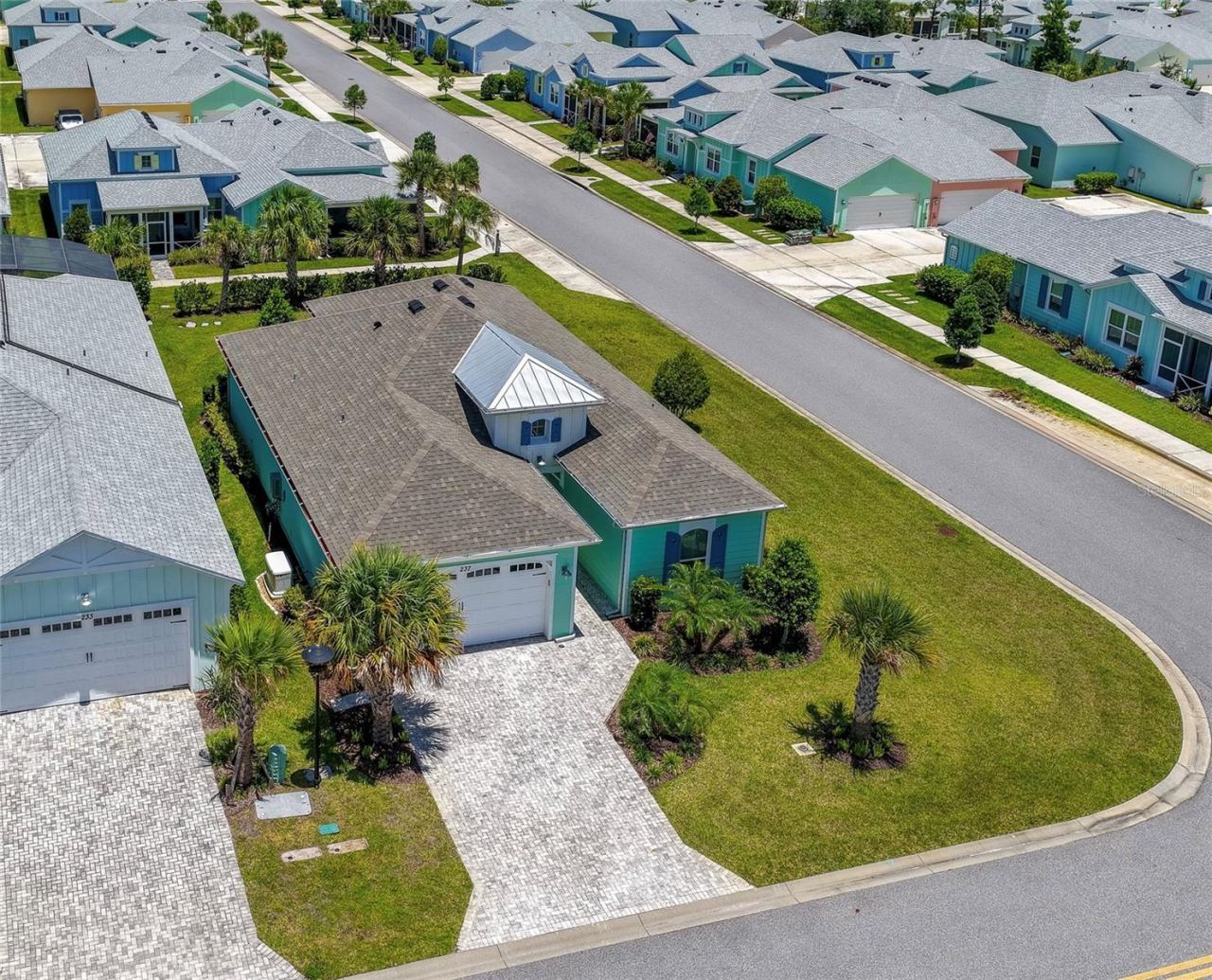 Photo 1 of 20: Beautiful aerial view of a coastal-style home featuring a paved driveway, palm trees, and a neatly manicured lawn. The surrounding neighborhood highlights charming, colorful houses along a quiet street.