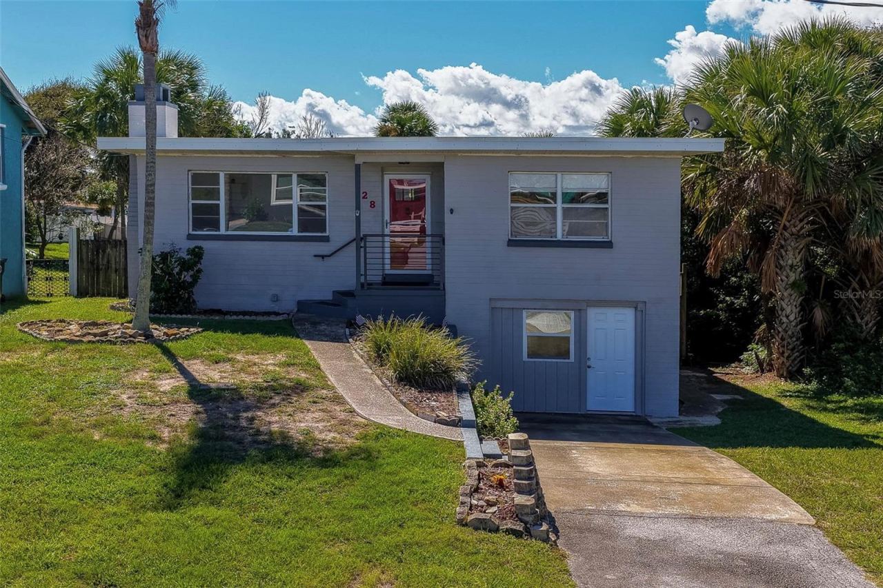 Photo 1 of 20: Exterior view of a coastal single-family home featuring a front driveway, grassy yard, and surrounding palm trees. The light gray facade offers elevated main entry steps and a lower ground-level entryway.