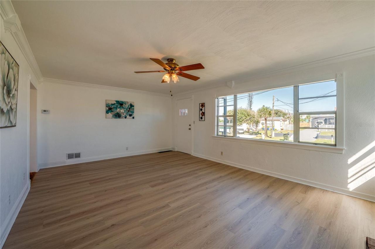 Photo 6 of 20: Bright living room highlighting smooth neutral walls, a ceiling fan with light fixtures, and a expansive front window overlooking the neighborhood. A side doorway connects seamlessly to the rest of the home's interior layout.