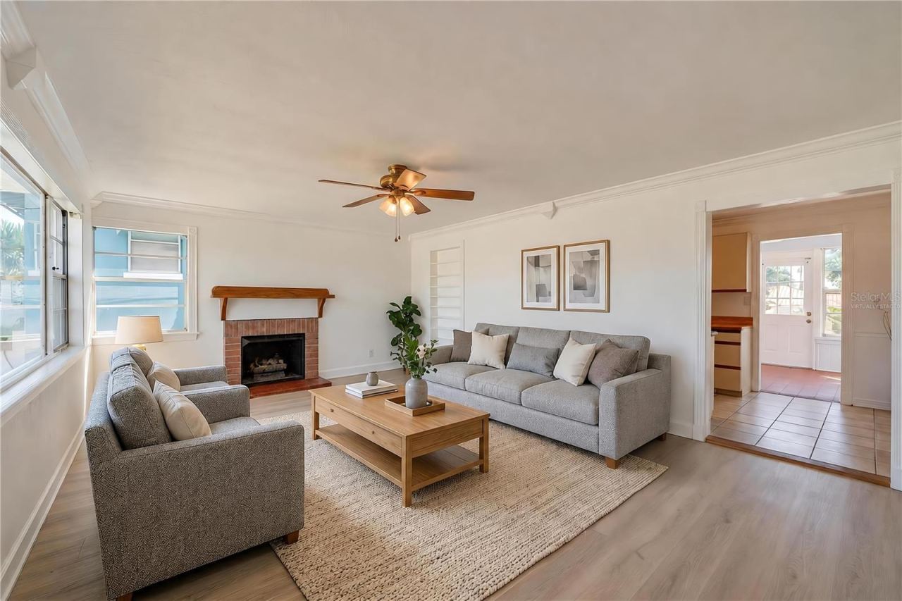 Photo 5 of 20: Virtually Staged - Spacious living area showcasing light LVP flooring, crown molding, and a charming brick fireplace with a wooden mantel. Large windows allow plenty of natural light, and an open doorway leads toward the kitchen and sunroom areas.
