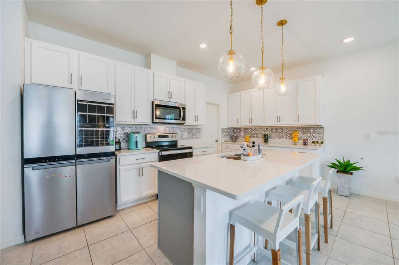 Photo 12 of 20: A close-up view of the kitchen showcasing a large island with a double sink, modern glass globe pendant lighting, and ample counter space. The white cabinetry is complemented by sleek stainless steel appliances and a geometric patterned backsplash.