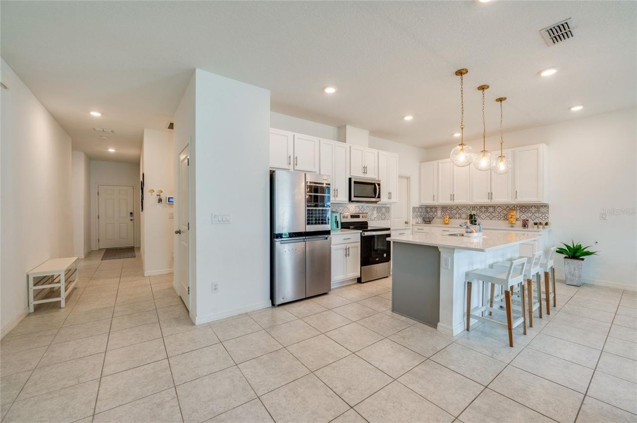Photo 7 of 20: Bright, open-concept kitchen featuring an island with a breakfast bar, white cabinetry, a decorative tile backsplash, and stainless steel appliances. The space flows seamlessly into a tiled hallway leading to the entryway.