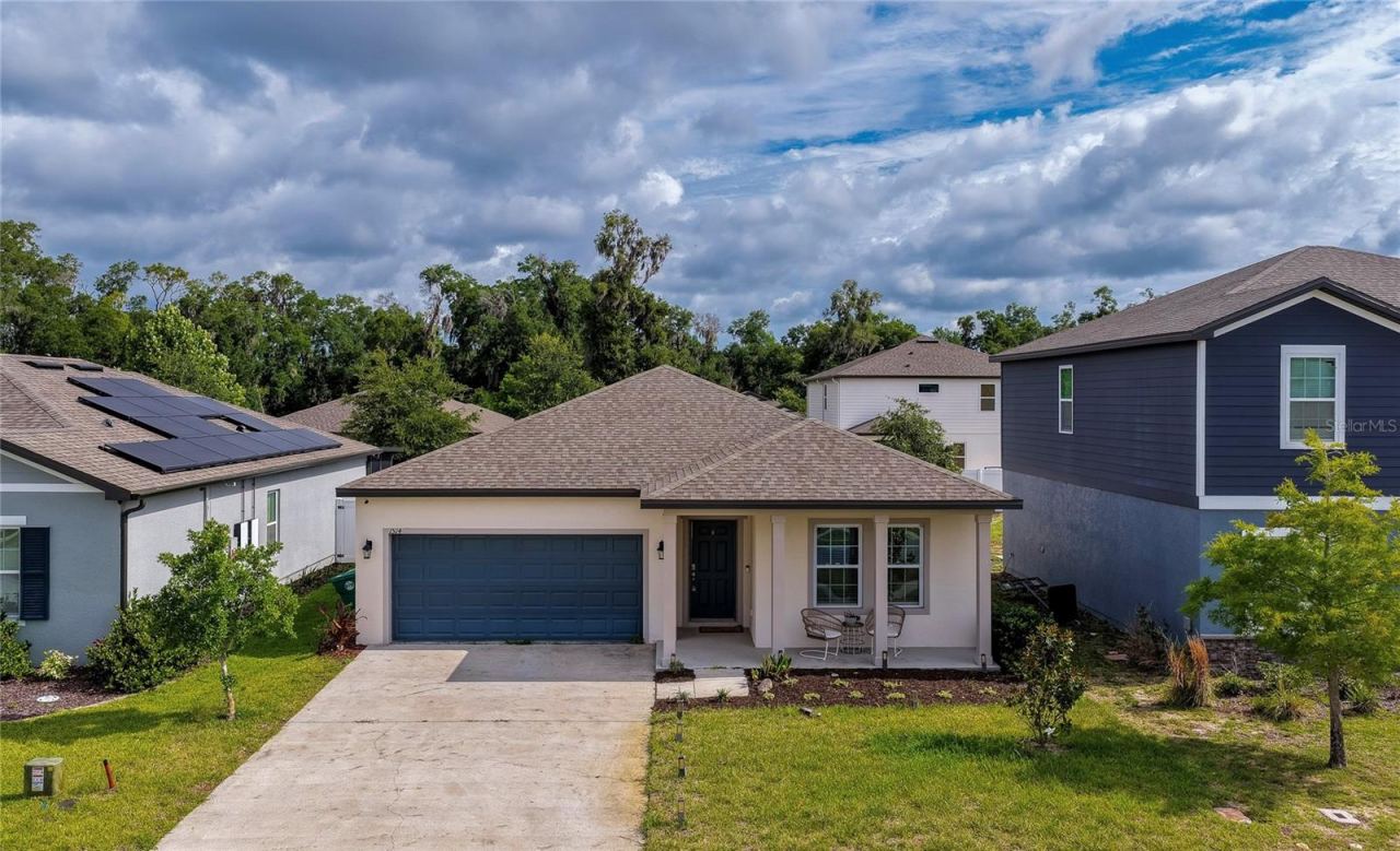 Photo 1 of 20: Charming single-story home featuring a two-car garage with a deep blue door, and a welcoming covered front entryway. The property has a concrete driveway and a neatly landscaped front lawn under a bright, cloud-filled sky.