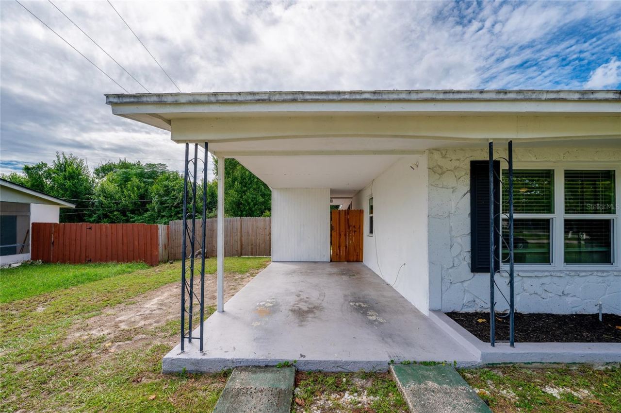Photo 4 of 20: Covered concrete carport with wrought-iron support posts, a textured exterior wall finish, and a gated side pathway.