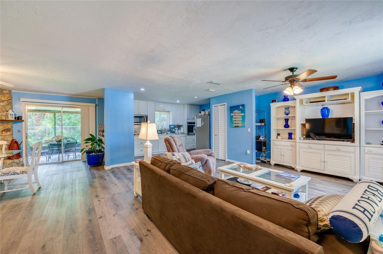 Photo 6 of 20: Expansive open living space featuring LVP flooring, bright blue walls, and a large built-in media center. Sliding glass doors open to the adjacent screened patio area.