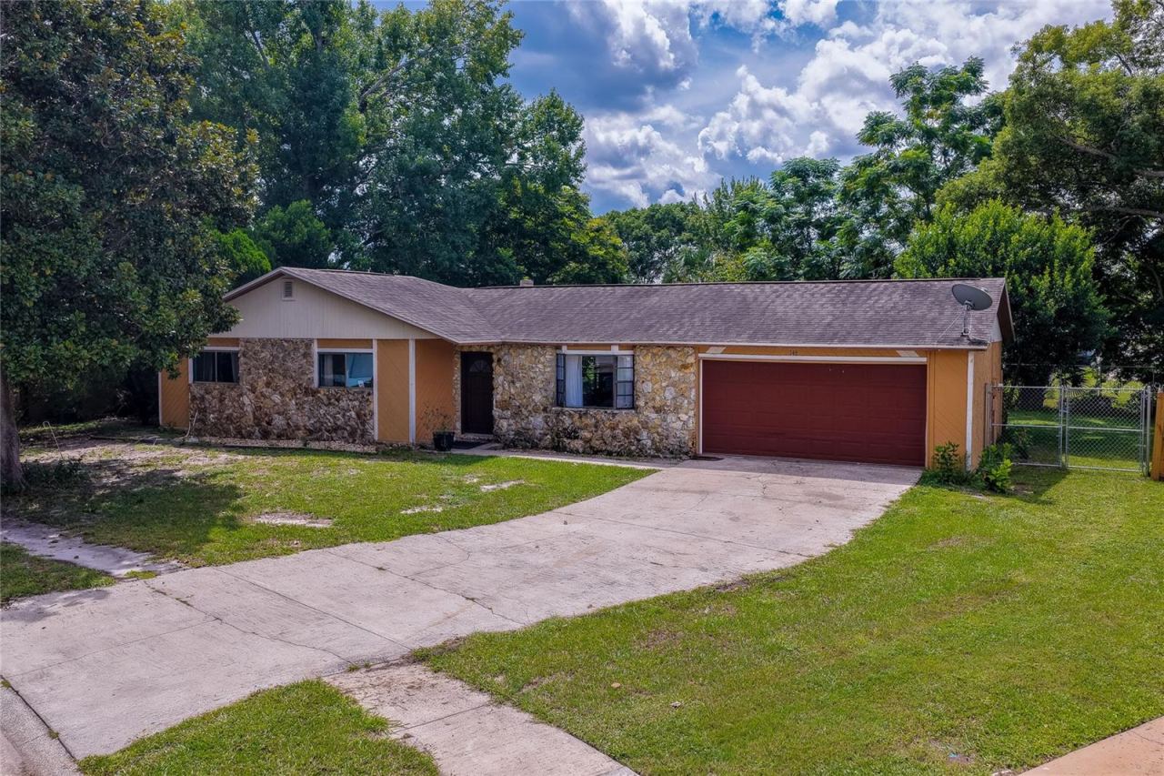 Photo 1 of 20: Charming single-story ranch home featuring a stone-accented facade, attached two-car garage, and an expansive paved driveway framed by lush green lawns and mature trees.