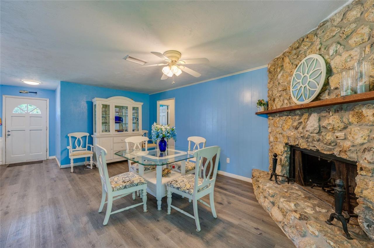 Photo 7 of 20: Open-concept living room anchored by a rustic stone fireplace, featuring light blue walls, LVP flooring, and a lighted ceiling fan.