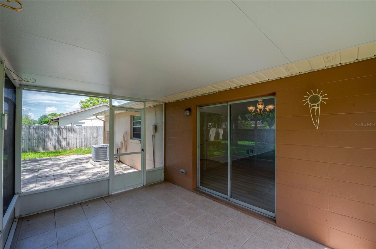 Photo 18 of 20: Covered patio area highlighted by durable tiled flooring, painted block walls, and a sliding glass door providing direct access into the home's interior. A screened door opens out toward an adjacent exterior paved patio space.