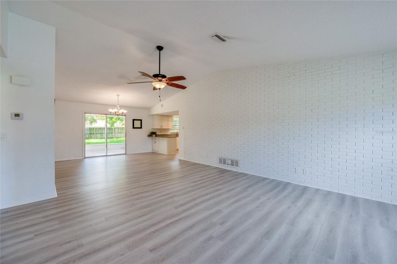 Photo 5 of 20: Spacious open-concept living area highlighting light luxury vinyl flooring, a painted white brick accent wall, and a ceiling fan. Sliding glass doors at the rear provide seamless access and views to the backyard patio.
