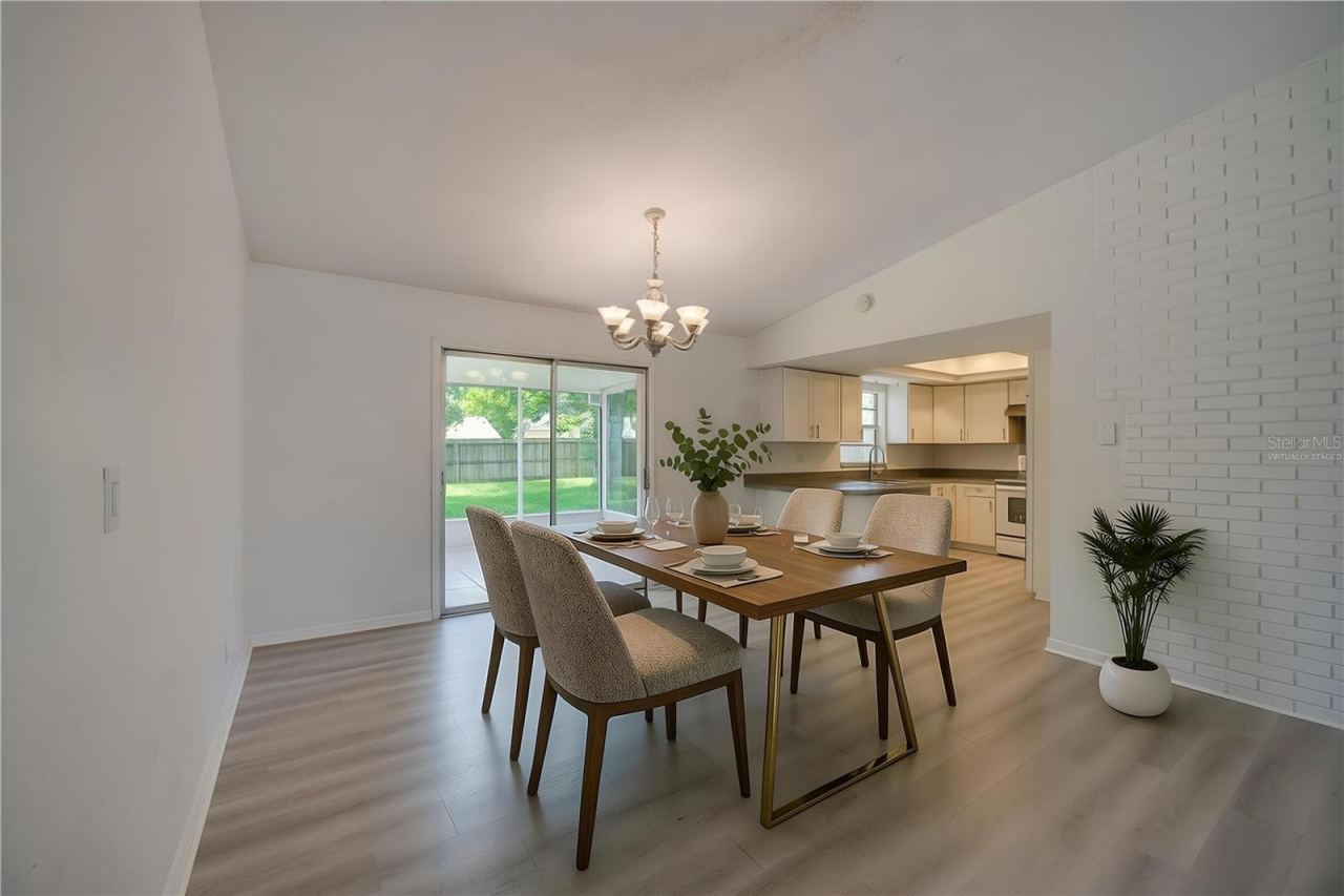Photo 12 of 20: Virtually Staged. Open dining and living space defined by a classic chandelier, light luxury vinyl flooring, and a white brick wall accent. Sliding glass doors open to a screened porch, filling the space with natural light.