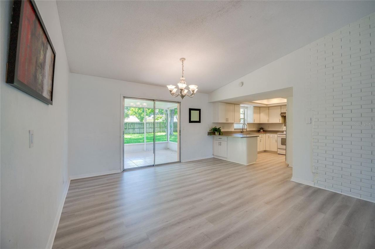 Photo 11 of 20: Open dining and living space defined by a classic chandelier, light luxury vinyl flooring, and a white brick wall accent. Sliding glass doors open to a screened porch, filling the space with natural light.