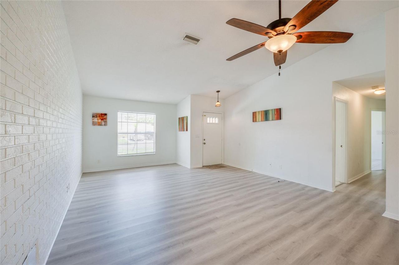 Photo 8 of 20: Welcoming foyer and main living room showcasing white walls, modern luxury vinyl flooring, and a ceiling fan with light fixture. A painted brick accent wall adds character to the functional entryway.