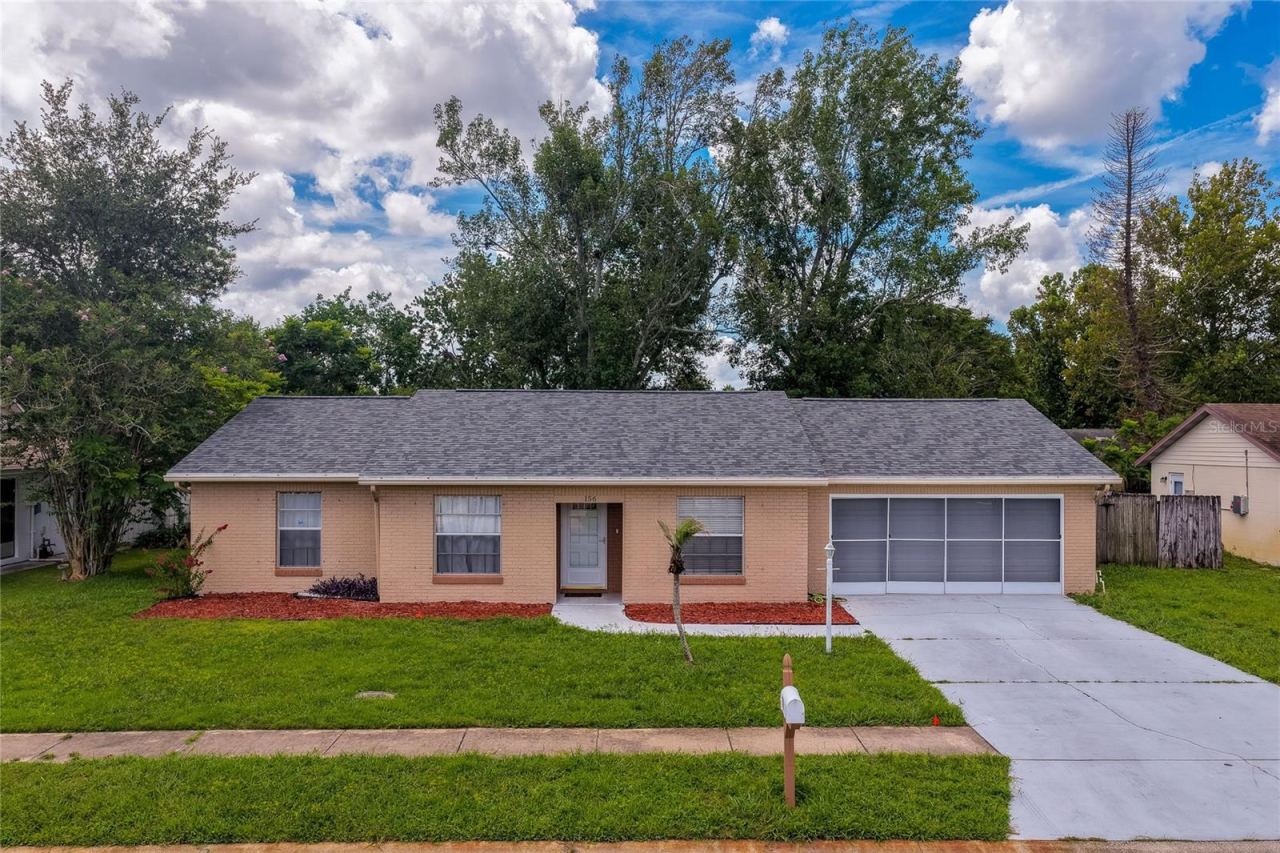 Photo 2 of 20: Charming single-story home featuring a spacious front lawn, concrete driveway, and screened garage enclosure. Lush surrounding trees and vibrant landscaping complete the inviting curb appeal.