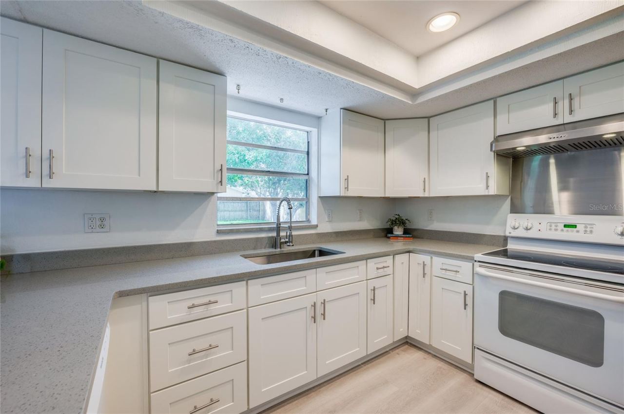 Photo 15 of 20: Well-lit kitchen area offering abundant white cabinetry, durable quartz countertops, and a window overlooking the backyard. A stainless steel range hood, smooth-top stove, and deep sink frame the efficient workflow.