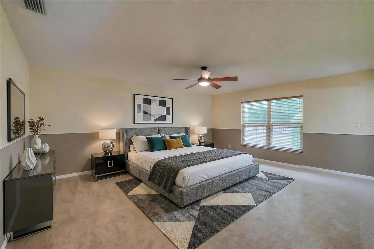 Photo 18 of 20: Virtually Staged - Spacious primary bedroom featuring soft carpeting, two-tone painted walls, a central ceiling fan, and a large window providing pleasant outdoor views.
